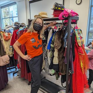 A woman in an orange shirt with a jack o lantern face stands by a rack of costumes