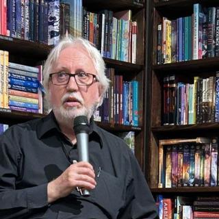 Perry Bulwer speaks into a microphone with a shelf of books behind him