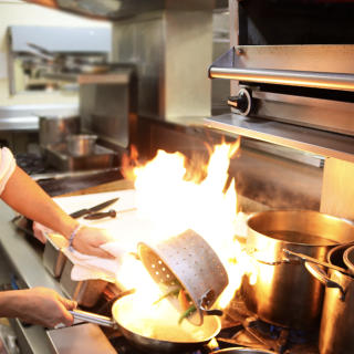 Culinary Student pouring vegetables into hot pan to sear them, creating short burst of flames in the pan