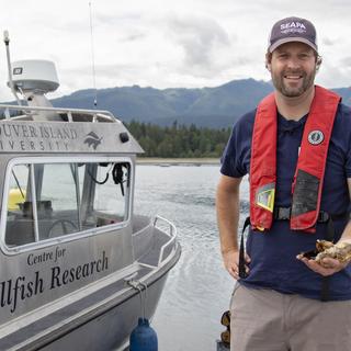 Dr. Timothy Green, VIU’s Canada Research Chair in Shellfish Health and Genomics stands on a dock next to a boat holding oysters in his hand.