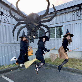 Three women pretend to fly on broomsticks with a large fake spider behind them.