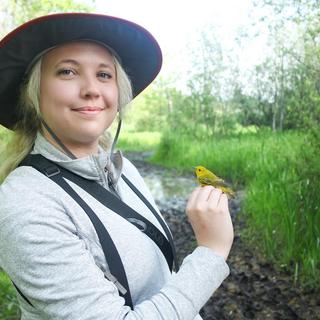 Chelsey Watts stands amongst bushes and trees holding a bird.