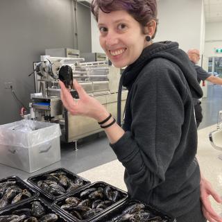 Melanie P. Loureiro stands in front of several bins of mussels and holds onel in her left hand.