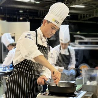 Ottis Crabbe wearing chef's hat and full cooking attire preparing a dish in a pot on a stove in an industrial kitchen during a competition.