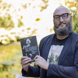 Dr. Theo Finigan, a VIU English Professor, holds up the book Atomic Blonde while standing in front of the VIU koi pond.