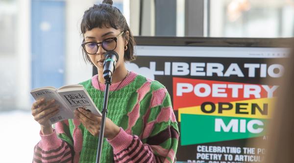 A woman wearing glasses and a pink and green sweater reads from a poetry book at a microphone