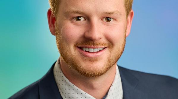 Mitch Turko wearing a suit and tie, smiling at the camera with a blue and green background.