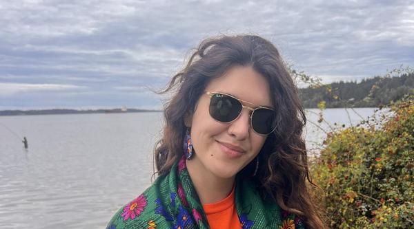 Alyona stands near a lake wearing a green shawl with pink, blue and yellow flowers over an Every Child Matters shirt.