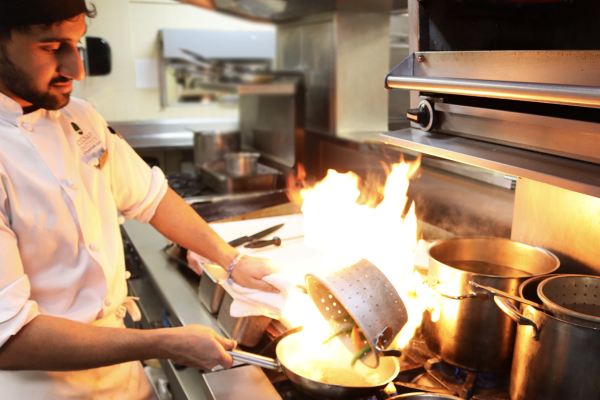 Culinary Student pouring vegetables into hot pan to sear them, creating short burst of flames in the pan