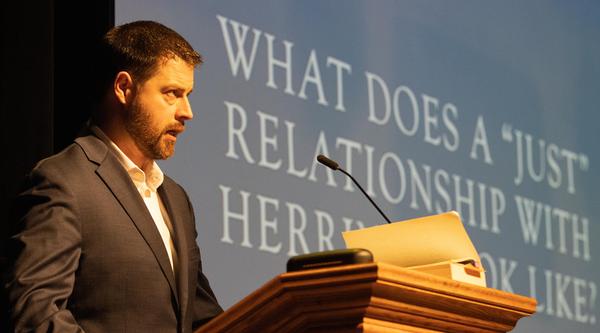 Man speaks at podium with text on a screen that reads: What does a just relationship with herring look like? 