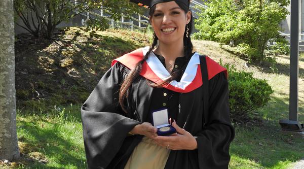 VIU Graduate, wearing convocation robes, Olivia Thomas is holding a medal while standing outside in front of a garden 