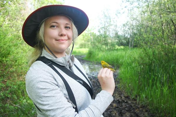 Chelsey Watts stands amongst bushes and trees holding a bird.