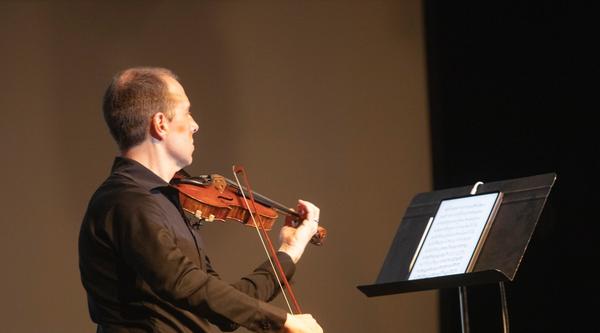 James Mark plays a violin while looking at a stand with sheet music on it.