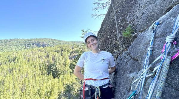 Sydney Solland stands on a ledge next to a rock face with climbing gear in front of her