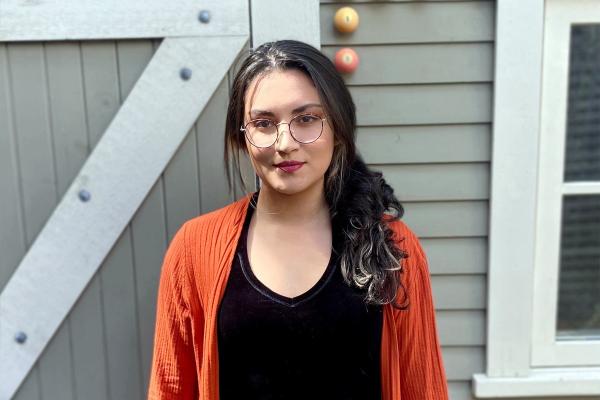 Sara Guzman stands in front of a building, wearing an orange sweater.