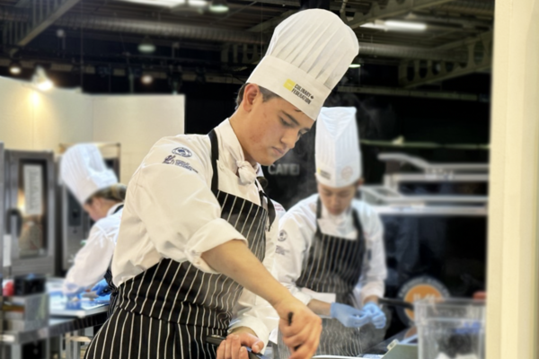 Ottis Crabbe wearing chef's hat and full cooking attire preparing a dish in a pot on a stove in an industrial kitchen during a competition.