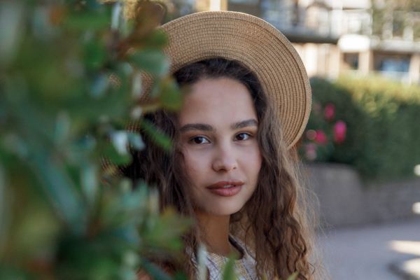 Anna Shynkarenko wearing a hat, half-hidden behind a bush and looking at the camera