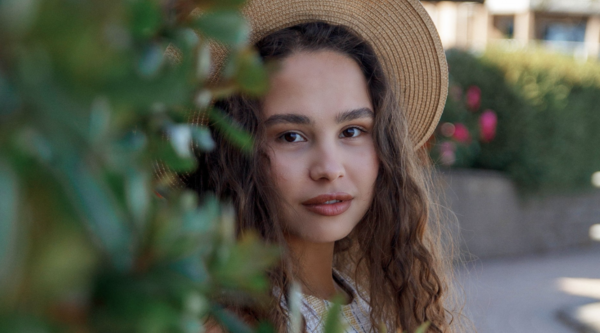 Anna Shynkarenko wearing a hat, half-hidden behind a bush and looking at the camera