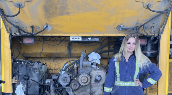 Madeline kozubal wearing mechanics coveralls standing in front of a school bus engine and smiling at the camera