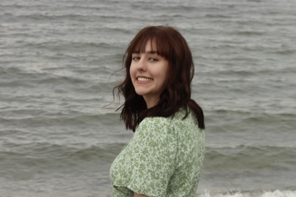 Paige Pierce standing in front of the ocean, wearing a green dress and turning back to smile at the camera