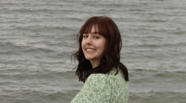 Paige Pierce standing in front of the ocean, wearing a green dress and turning back to smile at the camera