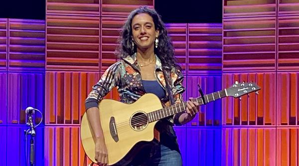 Sonnet L’Abbé sits on a stool on the Port Theatre's stage in front of a multi-coloured pink, blue and purple lighted background.