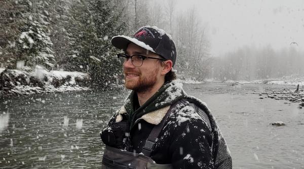 A young man clad in hipwaders is standing in a river with snowflakes falling around him