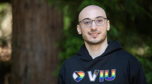 VIU grad and current employee Rayan Zeineddine wearing a VIU hoodie and glasses standing next to a tree outside and smiling at the camera