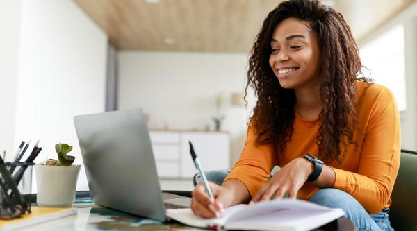 A woman takes notes and smiles while she looks at a laptop screen