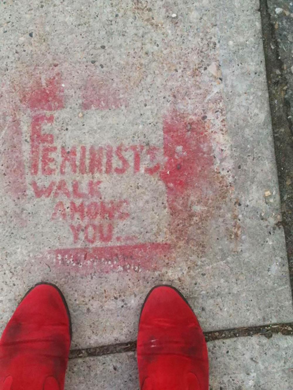 A light grey concrete sidewalk is spray painted with the phrase "Feminists Walk Among You."