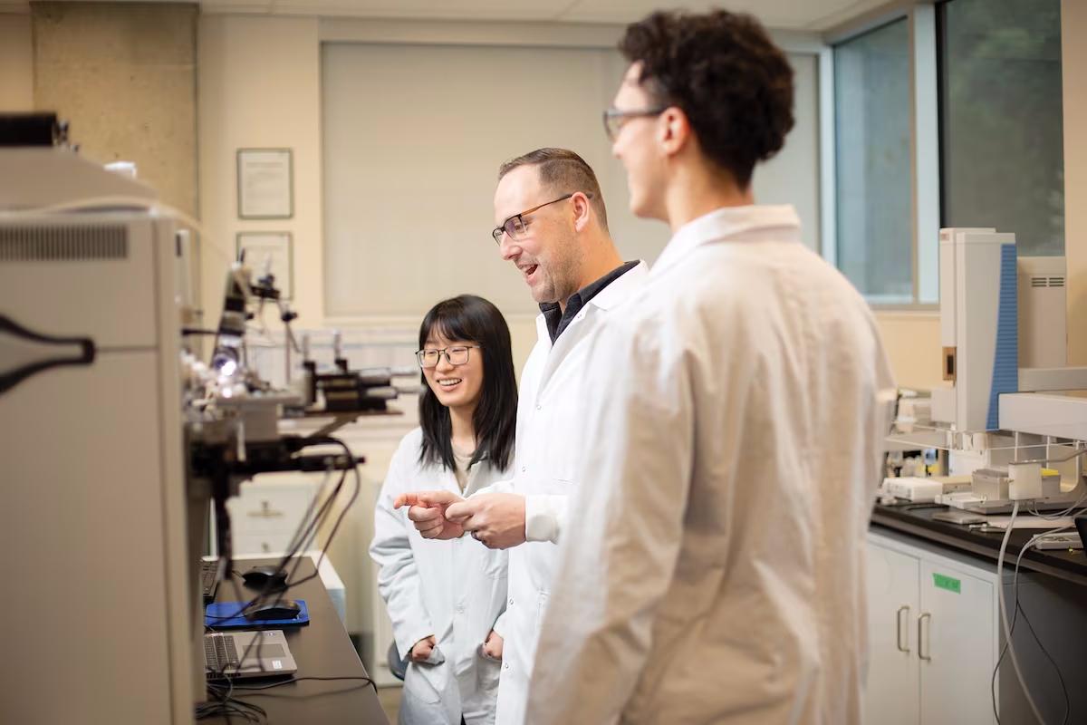 Dr. Kyle Duncan stands in front of a mass spectrometer with two students