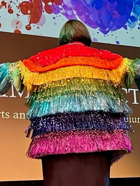 A woman displays a rainbow jacket with tassels, the view is of her back.