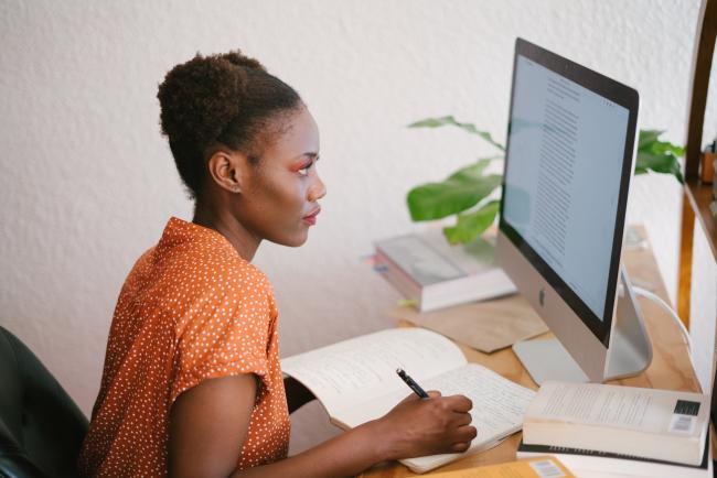 Female student at a desk taking notes and looking at a laptop screen