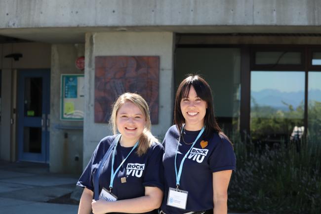 Two women wearing RockVIU shirts and lanyards smile in front of Building 310 at VIU's Nanaimo campus