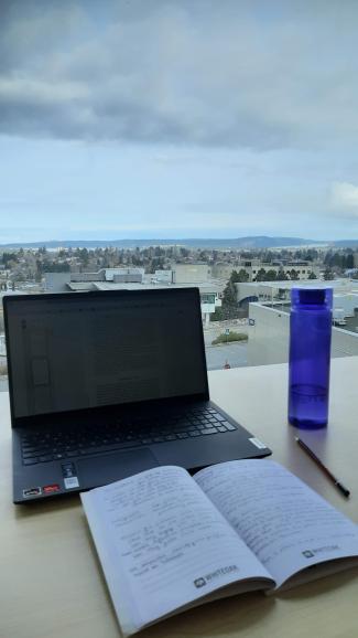 View of a laptop screen and notebook in the VIU library with a view of the city beyond